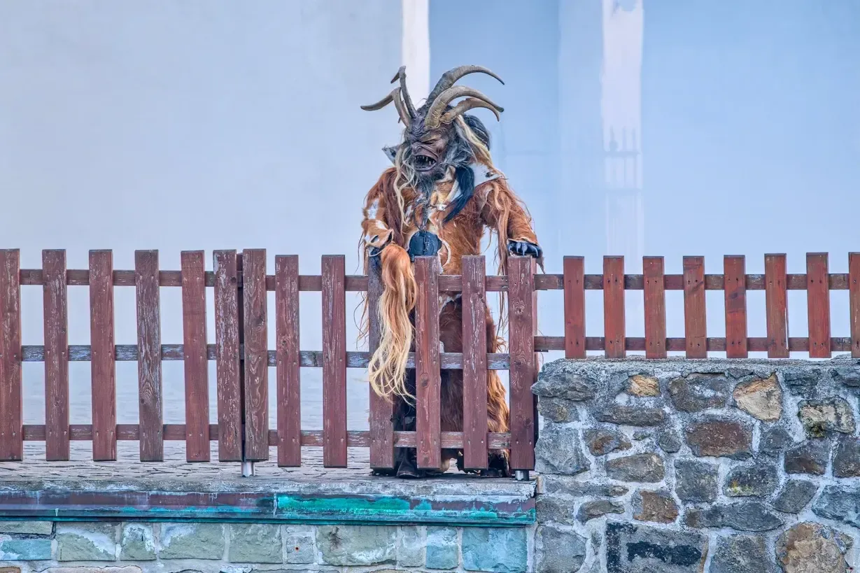 Krampus s rohy a kožešinou stojí na dřevěné palisádě nad kamennou hradbou